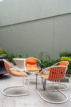 A patio area with three white metal chairs with orange cushions, a small round table, and a grey wall in the background. - Geko | Stylish Loft w/AC near Malecón de Barranco (Barranco)