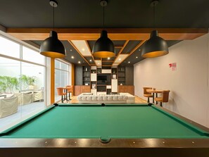 Une table de billard recouverte d’un drap vert, entourée de meubles en bois et éclairée par des lampes suspendues.