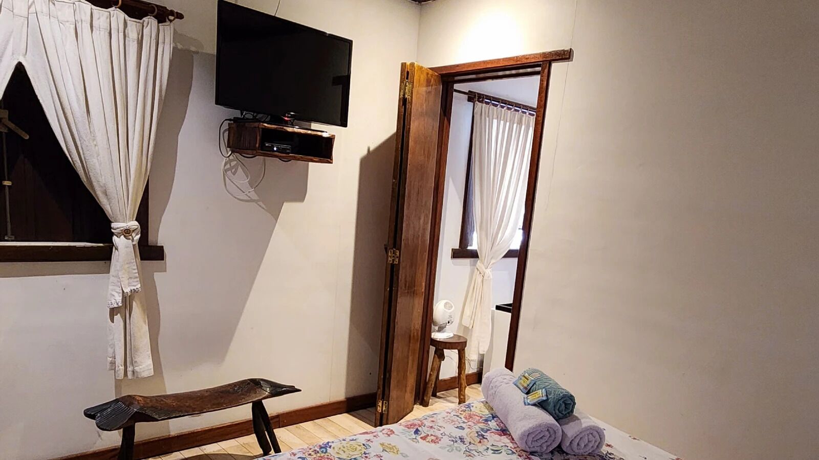Un dormitorio con cama, un banco de madera, un televisor montado en la pared y una puerta con cortinas.