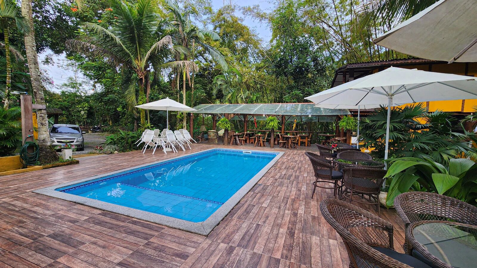 Piscina retangular com borda azulejada, rodeada por um deck de madeira. Há espreguiçadeiras brancas e mesas de vime com guarda-sóis.