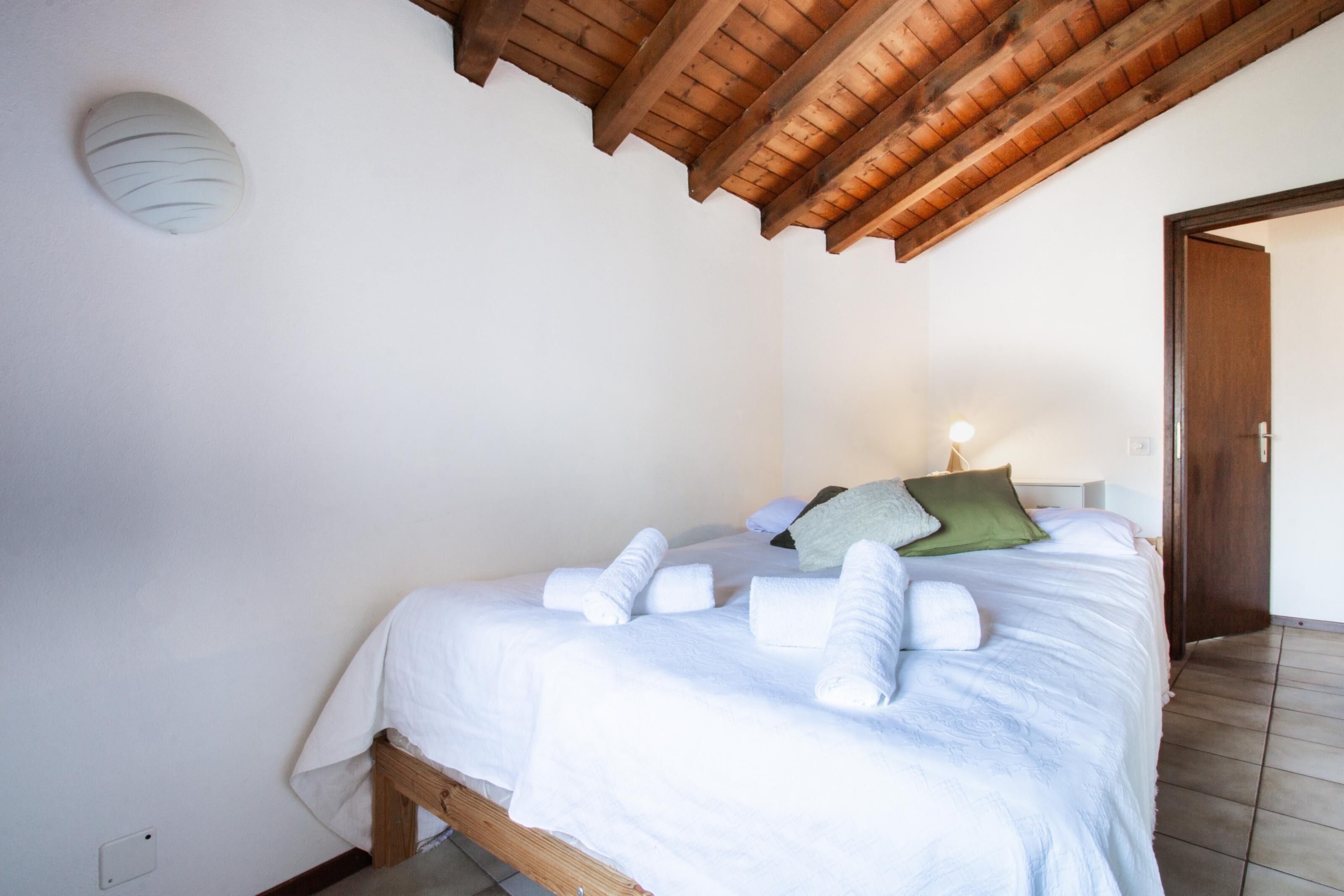 A bedroom with a wooden ceiling, a bed with white bedding and rolled towels, a green pillow, and a wall-mounted light fixture.