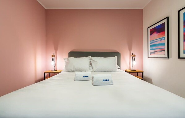 Un lit bien fait, avec du linge de lit blanc et des oreillers, deux tables de chevet avec des lampes, et des œuvres d’art encadrées accrochées au mur.