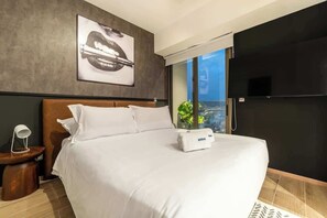 Ein modernes Hotelzimmer mit einem großen Bett, einem Nachttisch mit Lampe, einem gerahmten Kunstwerk an der Wand und einem großen Fenster mit Blick auf die Stadt.