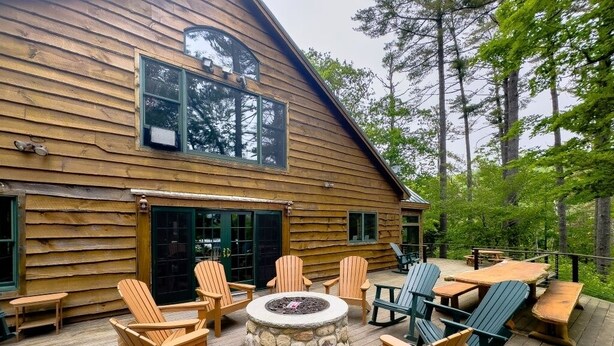 A wooden cabin with a deck, outdoor seating, and a stone fire pit.