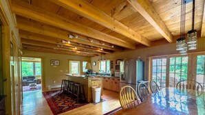 A spacious kitchen with wooden beams, a dining table with chairs, and a large window overlooking greenery.
