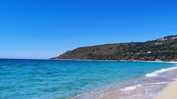 Une plage à l’eau turquoise et limpide, une rive sablonneuse et une colline parsemée de bâtiments.