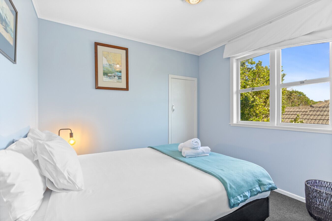 A bedroom with a bed, a window, a door, a wall-mounted light, and a framed picture.