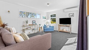 A modern living room with a grey sofa, a blue armchair, and a TV on a wooden stand. There are framed pictures on the wall and a small side table with a plant. - Cosy Retreat in Rothesay Bay| by Furnished Rentals (Auckland)