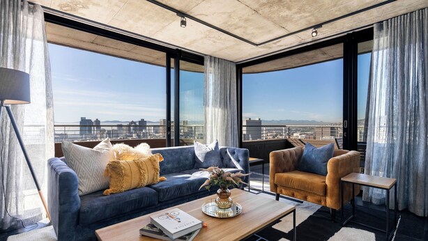 Un soggiorno moderno con un divano blu, una poltrona marrone e un tavolino da caffè. Ampie finestre offrono vista sulla città.
