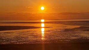 Een zonsondergang boven een strand, met de zon laag aan de horizon, waardoor een reflectie op het natte zand ontstaat.