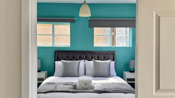 A neatly made bed with a grey headboard, white bedding, and a folded towel on the side. There are two bedside tables with lamps, and a window with a view of the outside.