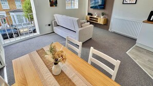 A modern living room with a sofa, a dining table with a vase of flowers, a TV mounted on a wooden panel, and a view of the street through large windows.