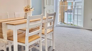 A dining area with a wooden table and white chairs, a vase with flowers, and a view of a residential street through a large window.