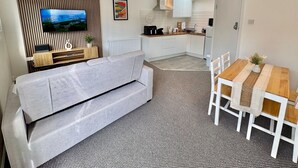A modern hotel room with a grey sofa, a wooden dining table with chairs, a kitchen area, and a TV mounted on a wall with a slatted design.