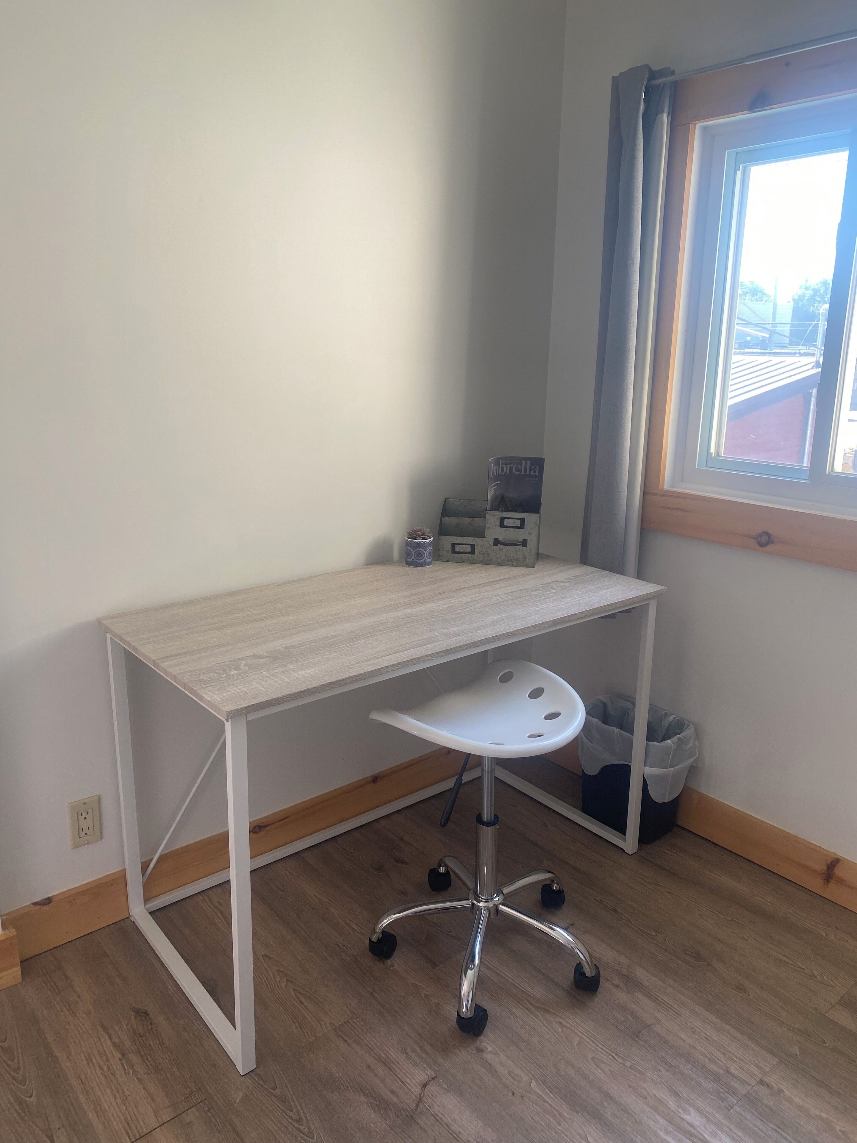 A small, neatly organized room with a light-colored desk, a white chair, and a window with a view of the outside.