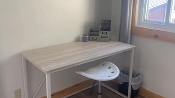 A small, neatly organized room with a light-colored desk, a white chair, and a window with a view of the outside.