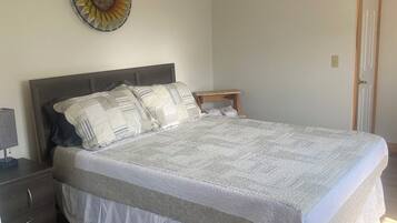 Un dormitorio con una cama grande, una mesita de noche, una pequeña mesa auxiliar de madera, un adorno en la pared y una puerta.