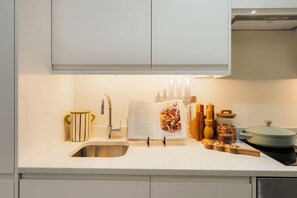 Une cuisine avec un plan de travail blanc, un évier en acier inoxydable, un livre de recettes, une planche à découper en bois, un bocal d’épices et un pot vert posé sur une plaque de cuisson à induction.