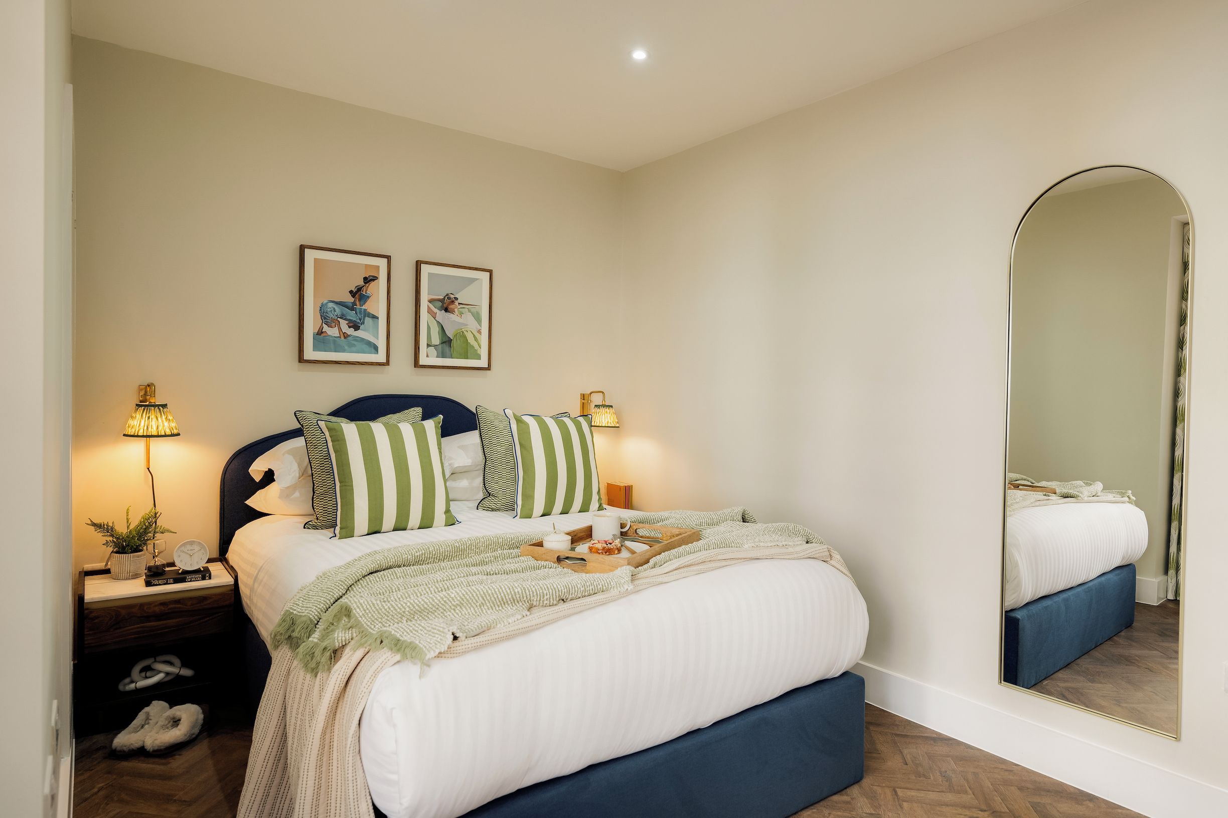 A neatly made bed with a headboard, a bedside table with a lamp, and a large mirror on the wall.