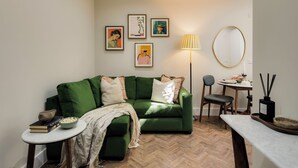 A green sofa with a white throw blanket, a small round table with a bowl of popcorn and books, a lamp, and a mirror on the wall. - Clifton Road Apartments | No.1 (Bristol)