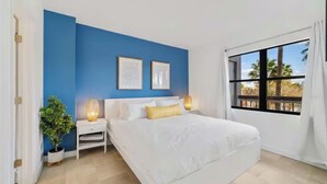 Een slaapkamer met een groot bed, twee nachtkastjes, een bijzettafeltje, een kamerplant en een raam met uitzicht op palmbomen.