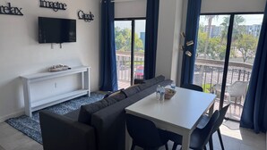 Een moderne woonkamer met een hoekbank, een eettafel met stoelen, een flatscreen televisie aan de muur en een groot raam met gordijnen.