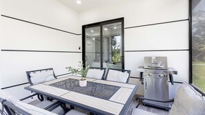 A modern outdoor dining area with a table and chairs, a grill, and a large window.