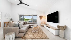 A modern living room with a sectional sofa, a TV, and a large window overlooking a garden. - Unit 4 Tuscany Shores (Marcoola)