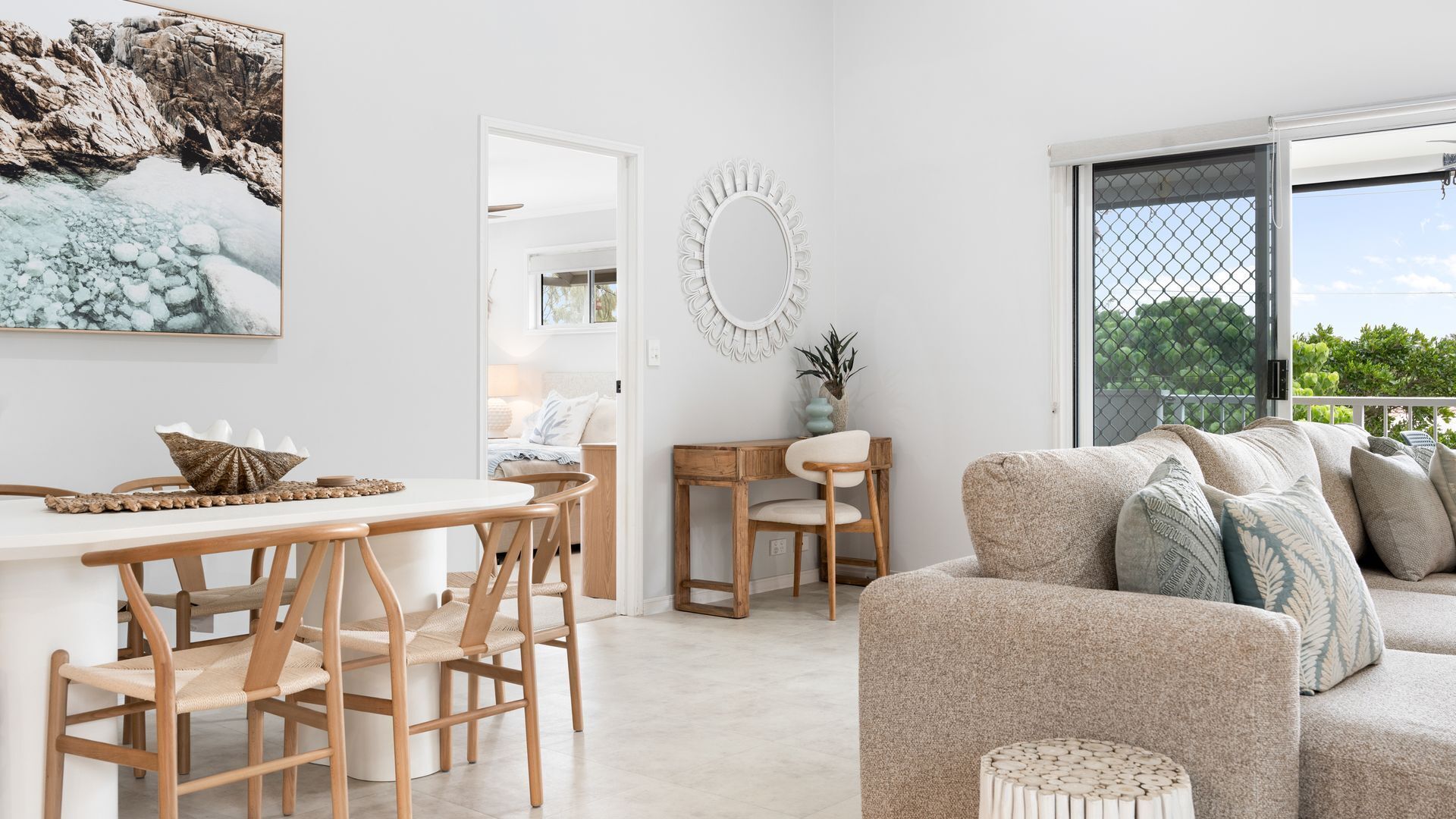 A modern living room with a sofa, a dining area with chairs and a table, a decorative mirror, and a view through a large window.