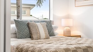 A bedroom with a large bed, multiple pillows, a bedside lamp, and a view of a building through a window. - Unit 4 Tuscany Shores (Marcoola)