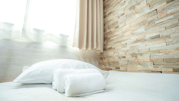 A neatly made bed with white linens and pillows, a stone-textured wall, and a window with curtains. - Japanese-style family house in the western of Tokyo, DP Oyamamachi　 (町田市小山町)