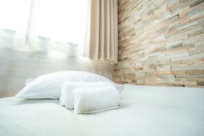 Un lit bien fait, avec du linge de lit blanc et des oreillers, un mur à la texture de pierre et une fenêtre avec des rideaux.