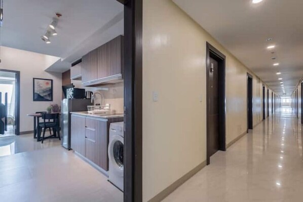 A modern hotel corridor with a kitchen area on the left, featuring cabinets, a microwave, and a washing machine.