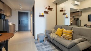 A modern living room with a grey sofa, wooden side tables, and a small round ottoman. There is a kitchen area with wooden cabinets and a microwave.