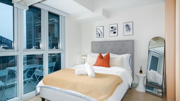 A modern hotel room with a large bed, a bedside table, a mirror, and a view of the outside through large windows. - Luxury 1BR at BGC Uptown with Free Parking (Taguig City)
