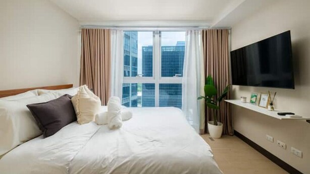 A hotel room with a large bed, a TV mounted on the wall, and a window with curtains. - New & Modern Uptown BGC 1BR 4pax (Taguig City)