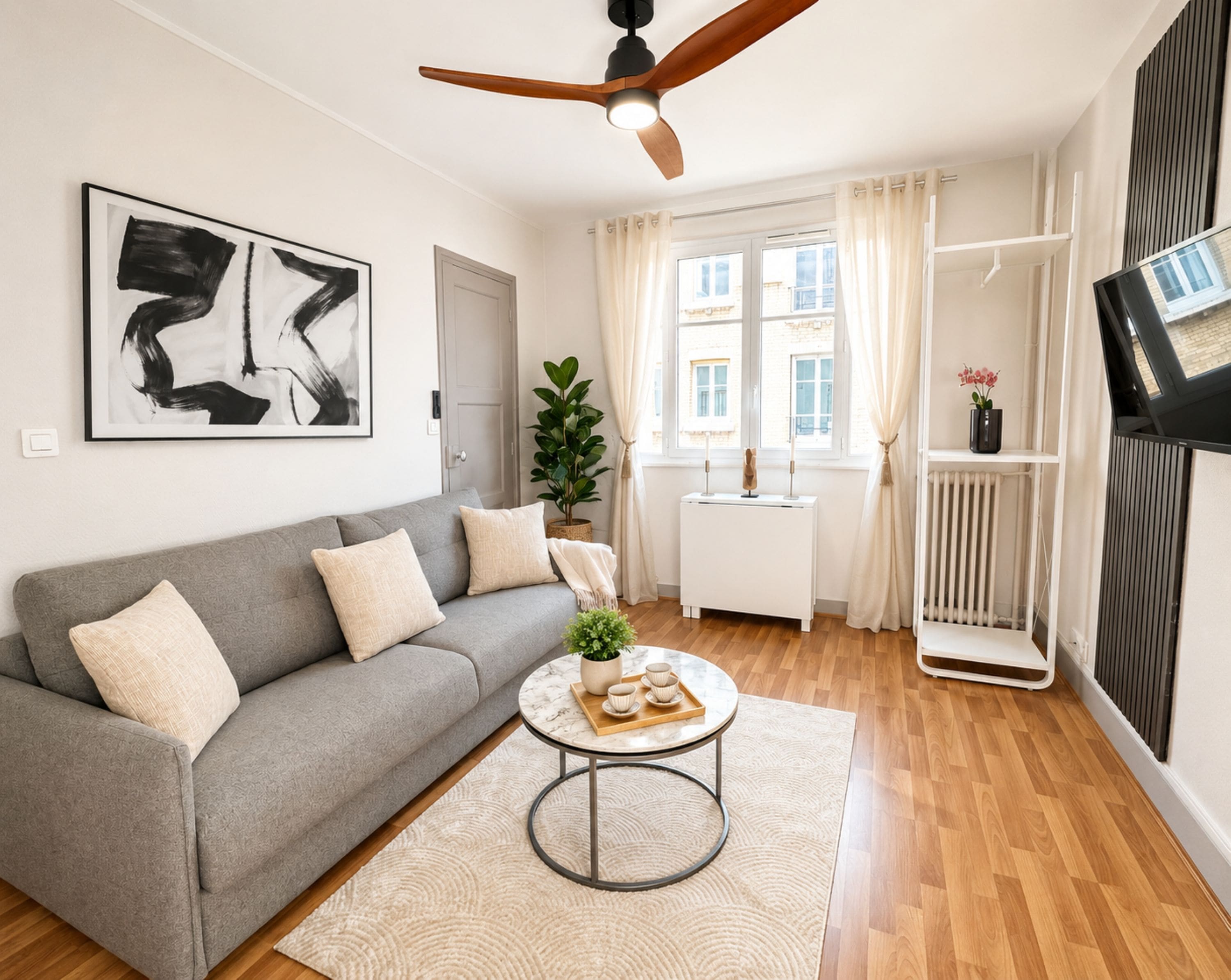 Ein modernes Wohnzimmer mit einem grauen Sofa, einem runden Couchtisch, einem Deckenventilator und einem großen Fenster mit Vorhängen.