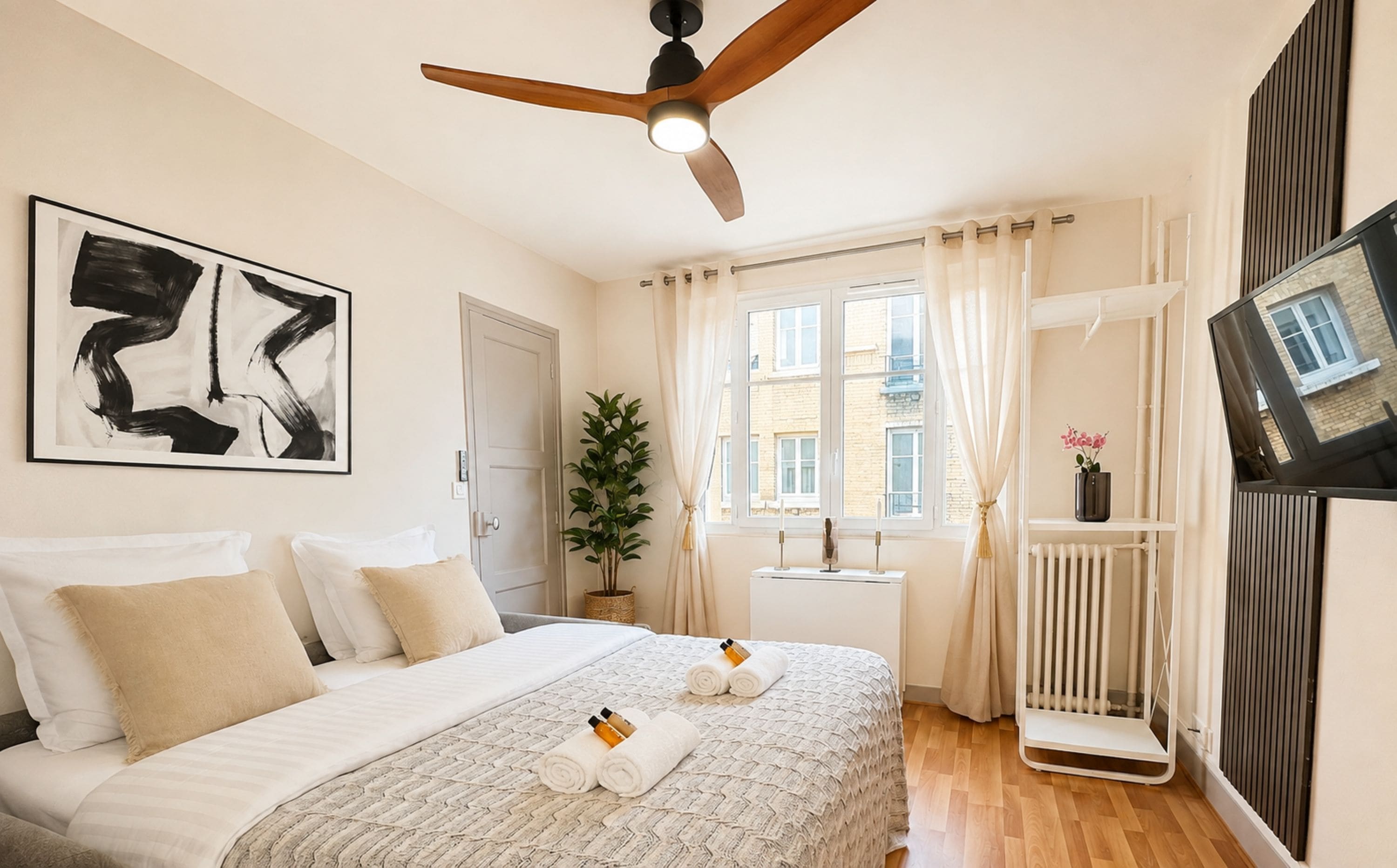 Een hotelkamer met een bed, een plafondventilator, een raam met gordijnen, een radiator en een aan de muur bevestigde televisie.