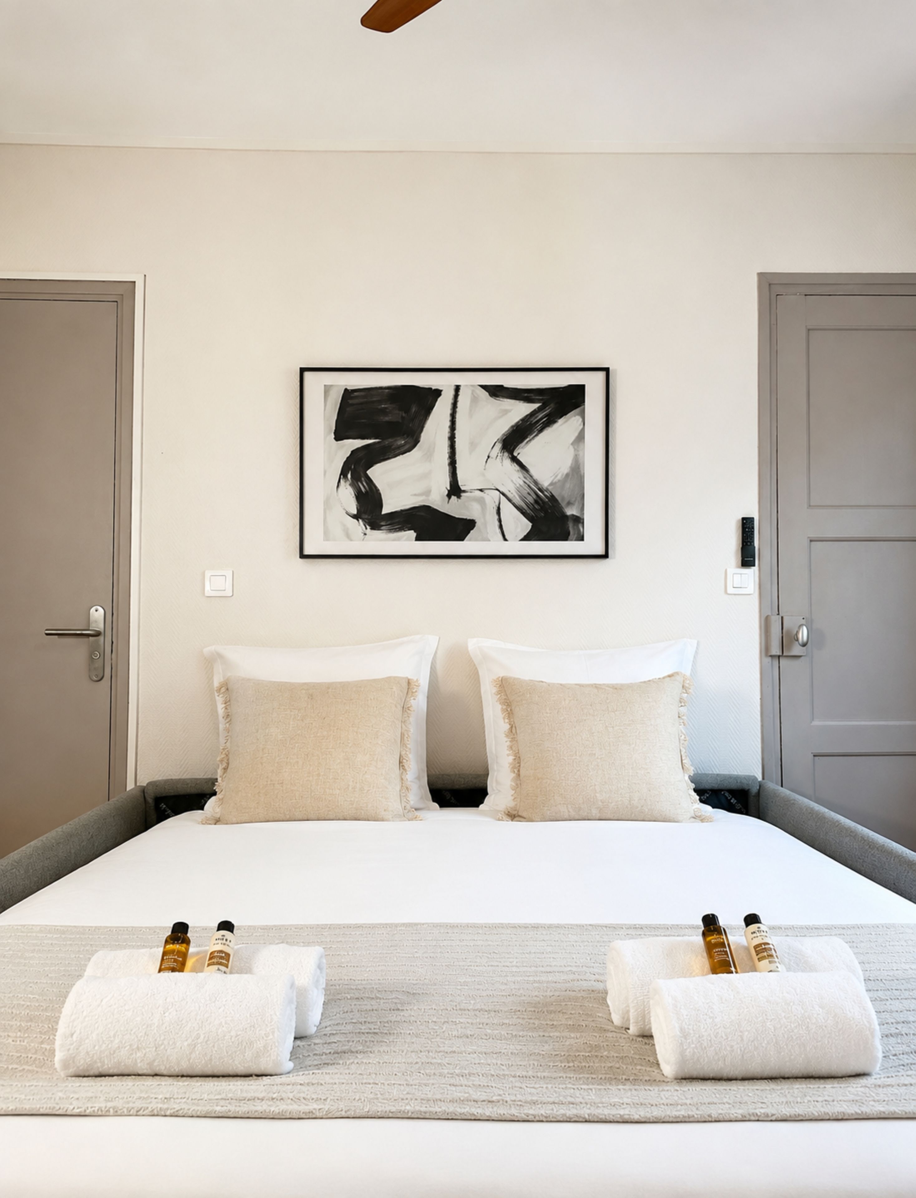 Habitación de hotel con una cama grande, dos almohadas, una obra de arte abstracta enmarcada, una puerta y toallas con botellas sobre la cama.