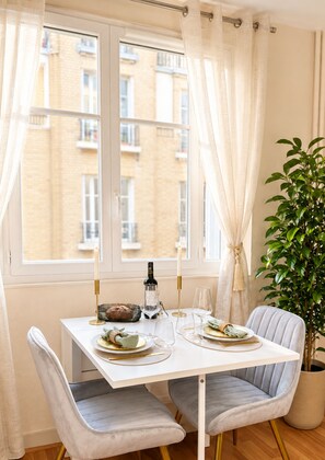 Zona pranzo con tavolo apparecchiato per due, una pianta in vaso e vista su un edificio attraverso una finestra.