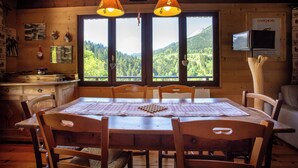 Une table à manger en bois, recouverte d’une nappe à carreaux, est entourée de chaises en bois. On aperçoit, au fond, une fenêtre donnant sur un paysage montagneux.