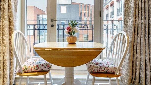 Un comedor con una mesa redonda de madera y dos sillas blancas con cojines estampados. Un jarrón con flores está sobre la mesa.