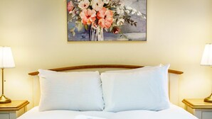 Una cama bien hecha con sábanas blancas y dos almohadas, con mesitas de noche a cada lado con lámparas. Sobre la cama cuelga un cuadro con flores.