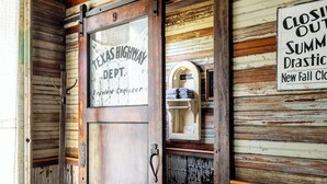 Una habitación rústica con una puerta de madera que indica “Ingeniero Residente del Departamento de Carreteras de Texas”, una cama con cabecera decorativa y un letrero que dice “Liquidación de Verano.”