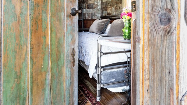 Una habitación rústica con una puerta de madera, una cama con ropa de cama blanca y una pared de piedra.