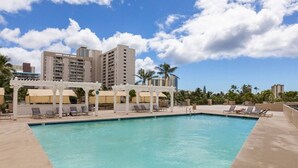 A swimming pool with lounge chairs and umbrellas, surrounded by palm trees and tall buildings.