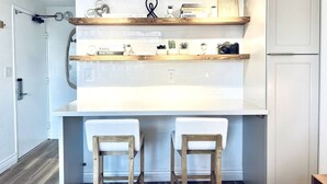 A kitchen with white cabinets, a white countertop, two wooden stools, and shelves with decorative items and a sign.
