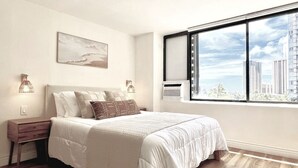 A bedroom with a large bed, a nightstand, a wall-mounted lamp, and a window with a city view.