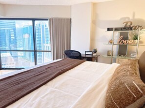 Une chambre d’hôtel avec un grand lit, un bureau, une chaise, une fenêtre donnant sur la ville et une étagère décorative ornée de différents objets.