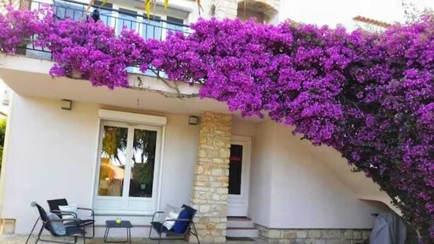 Un bâtiment à la façade en pierre, un balcon orné de bougainvilliers violets et deux fauteuils confortables à l’extérieur.